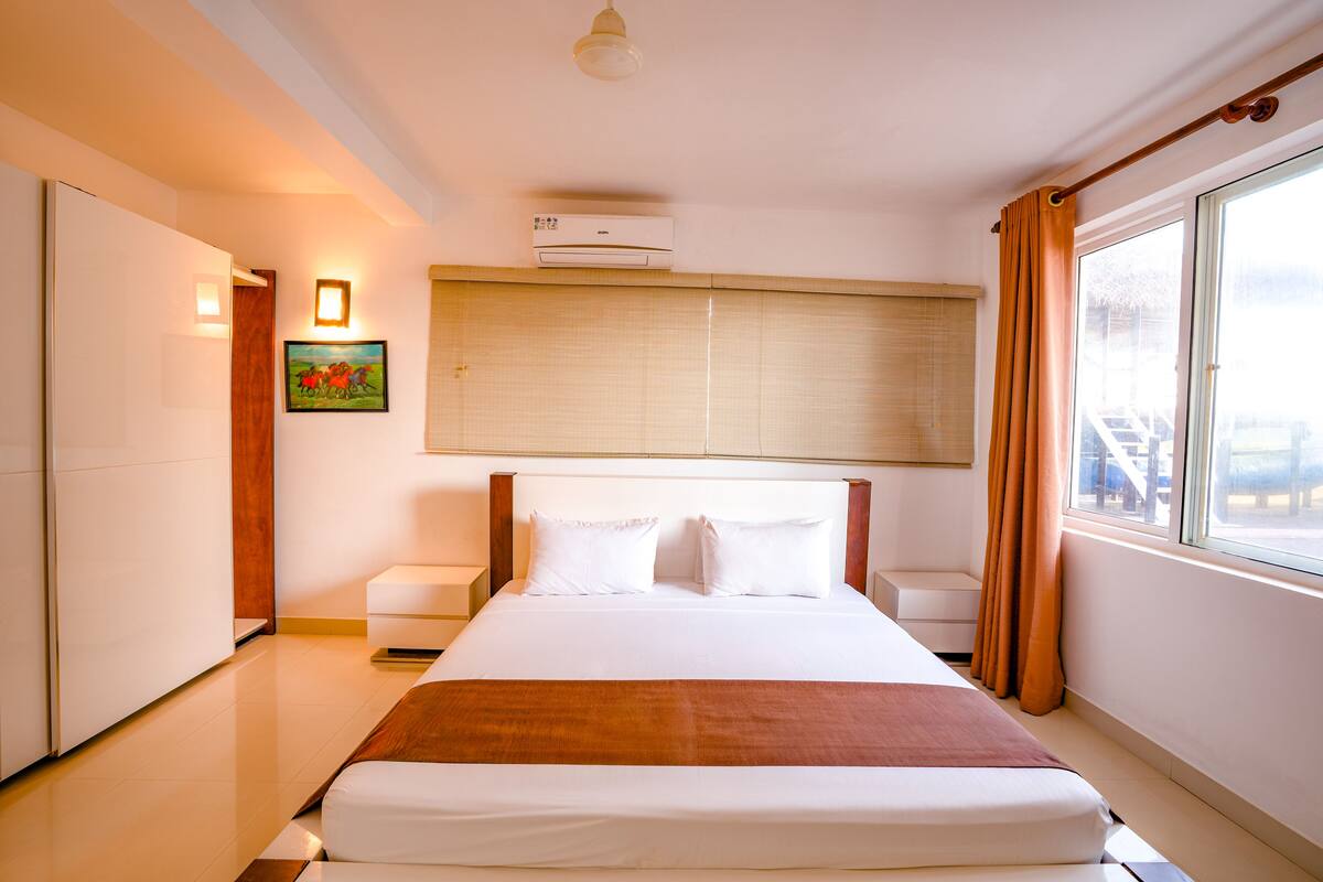 A hotel room with a large bed, a bedside table, a nightstand, a window with curtains, a wall-mounted air conditioner, and a painting on the wall.