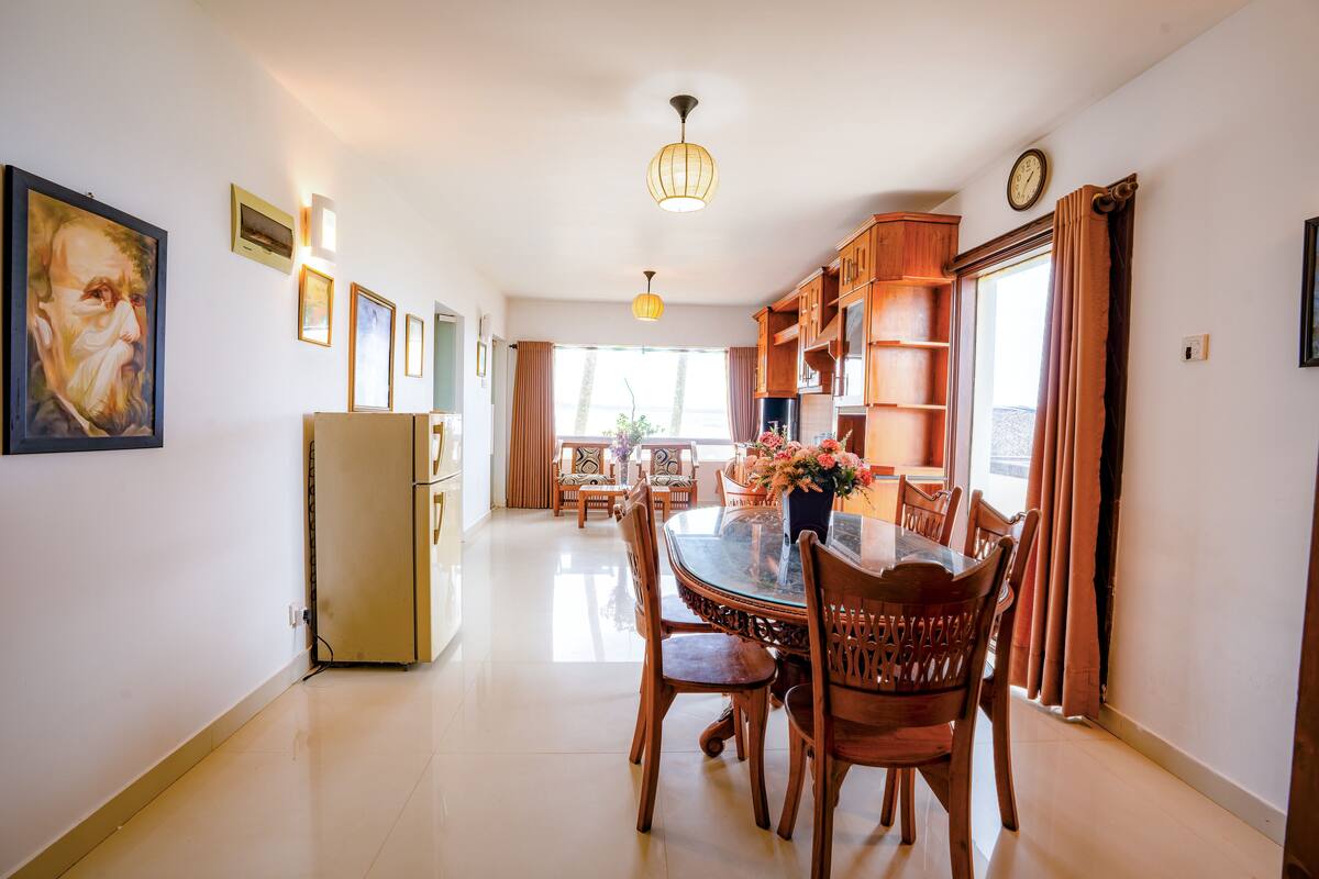 A dining area with a glass-top table, wooden chairs, and a chandelier. There is a refrigerator and a clock on the wall.