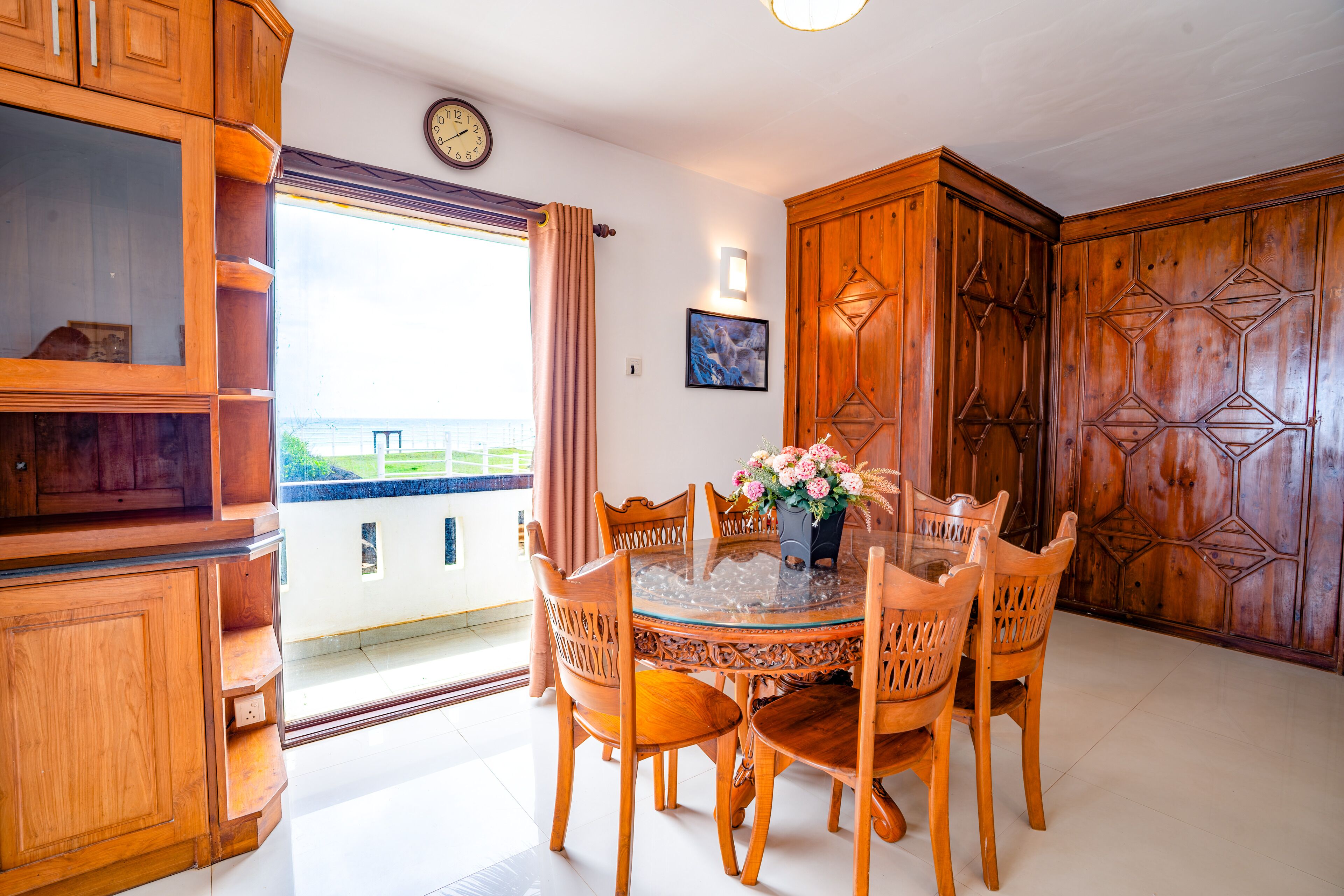 A dining area with a round glass table surrounded by wooden chairs, a wooden cabinet with a geometric design, and a clock on the wall.
