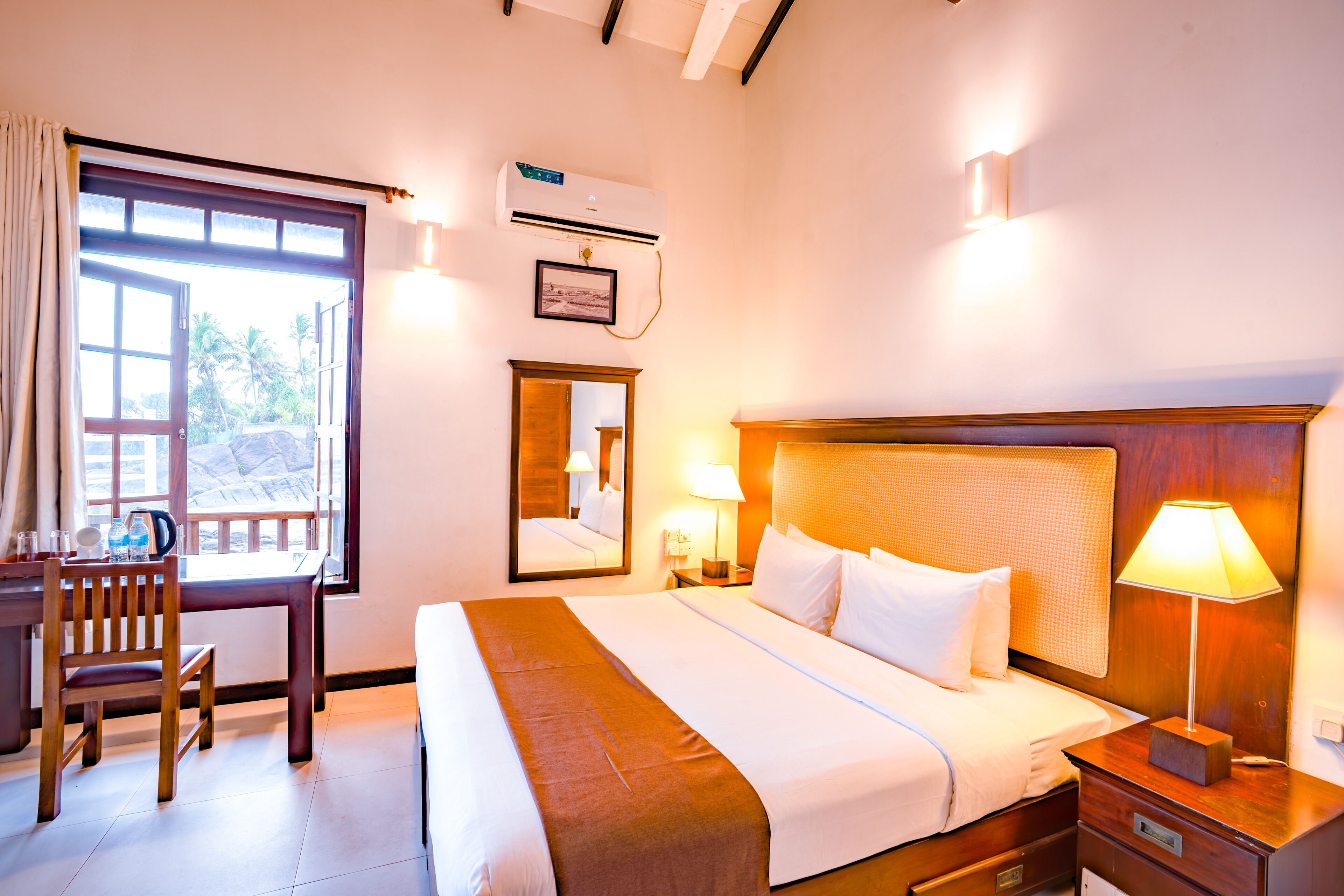 Habitación de hotel con cama, mesita de noche, lámpara, escritorio con silla y ventana con cortinas.