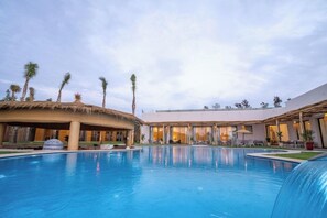 A swimming pool with a thatched roof structure, palm trees, and a modern building with large windows.