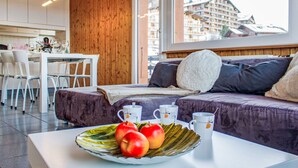 A modern room with a wooden ceiling, a sofa with cushions, a dining area with chairs, and a window with a view of buildings. - Bietschorn 20 by Interhome (Nendaz)