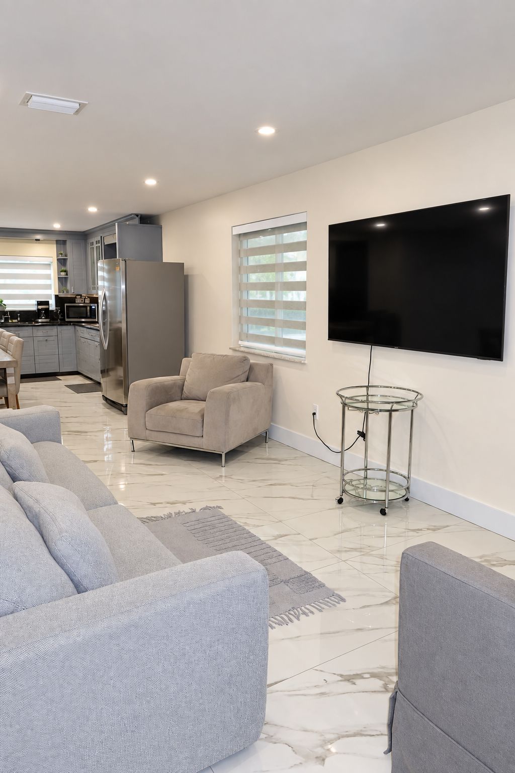 A living room with a sofa, a TV mounted on the wall, and a kitchen in the background.
