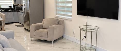A living room with a sofa, a TV mounted on the wall, and a kitchen in the background.