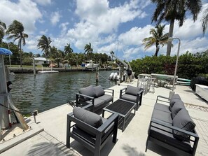 A waterfront patio with outdoor furniture, a pool, and palm trees.