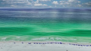 A beach with clear turquoise water, a sandy shore, and a row of blue umbrellas. - Majestic Sands | Free Golf Cart & Beach Chairs | Gorgeous Private Pool & Courtyard | 2 PM Check In! | Clubhouse Amenities! Pickleball, Gym & Hot Tub! (Destin)