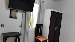 Quarto com televisão de tela plana na parede, uma mesinha, uma cadeira com almofada, guarda-roupa e janela com cortinas.