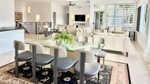 A spacious living room with a glass dining table, multiple chairs, a sofa, and a TV. There are ceiling fans, a staircase, and a large window with blinds.