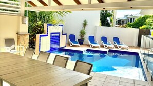 A covered outdoor area with a pool, lounge chairs, and a dining table.