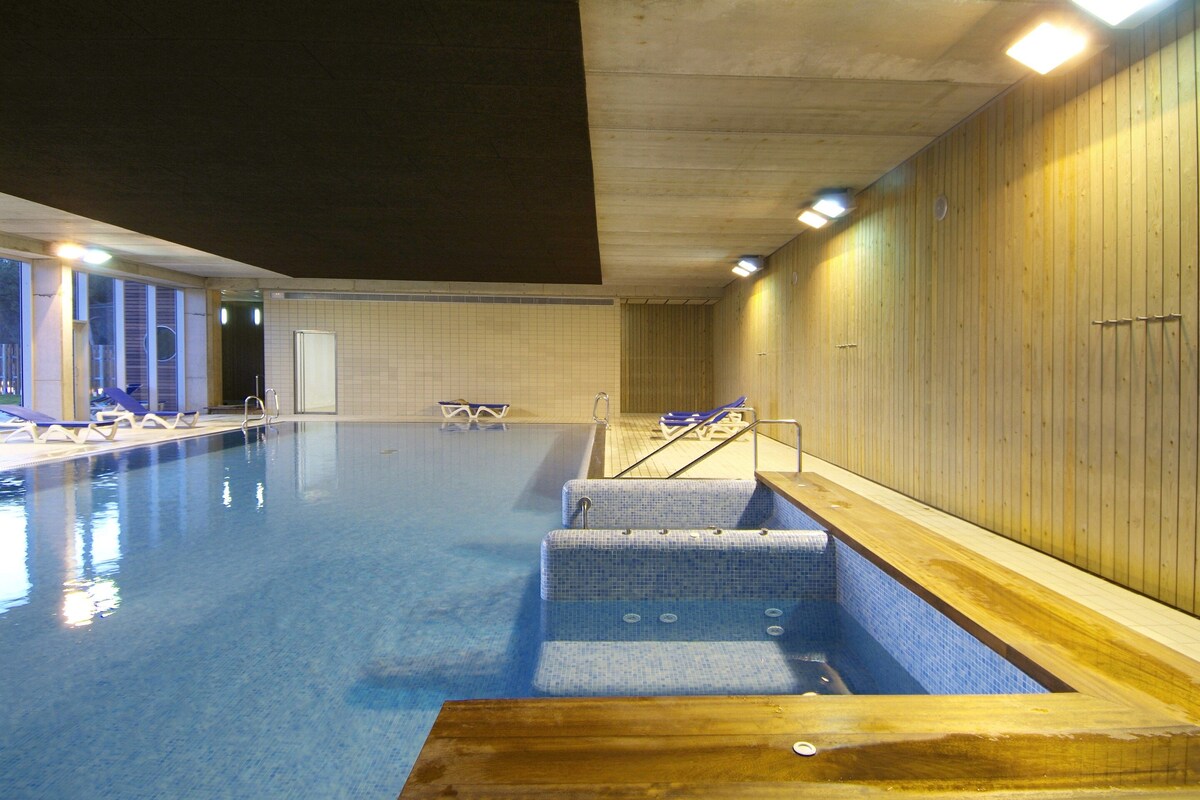 An indoor swimming pool with clear water, steps leading into the pool, and lounge chairs along the side.