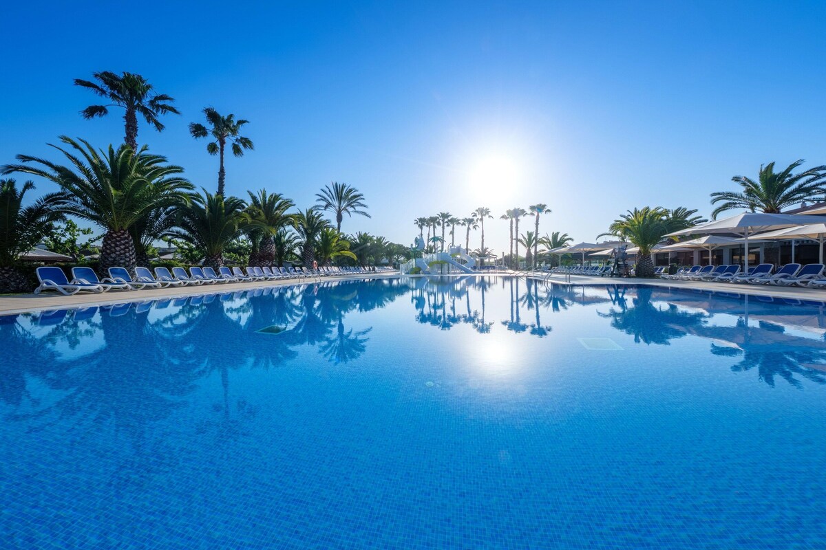 A clear blue pool surrounded by lounge chairs and palm trees, with a bright sun in the background.