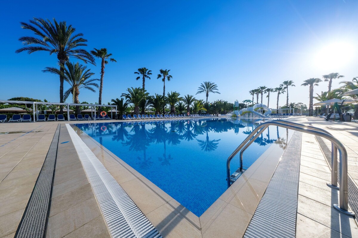 A swimming pool with clear blue water, surrounded by palm trees and lounge chairs.