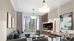 A modern living room with a large flat-screen TV, grey sofas, a dining table, and a view of the cityscape through large windows.