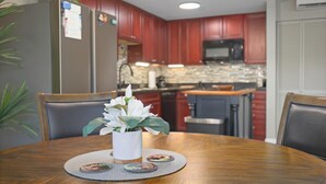 Un comedor con una mesa de madera, sillas, un jarrón con flores y una cocina al fondo.
