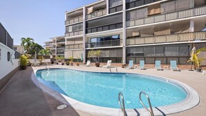 Piscina con sillones de descanso y plantas en macetas, rodeada por un edificio de varios pisos con balcones.
