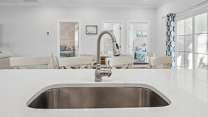 Une cuisine moderne dotée d’un évier en acier inoxydable, de plans de travail blancs et d’un coin repas avec des chaises et une table blanches.