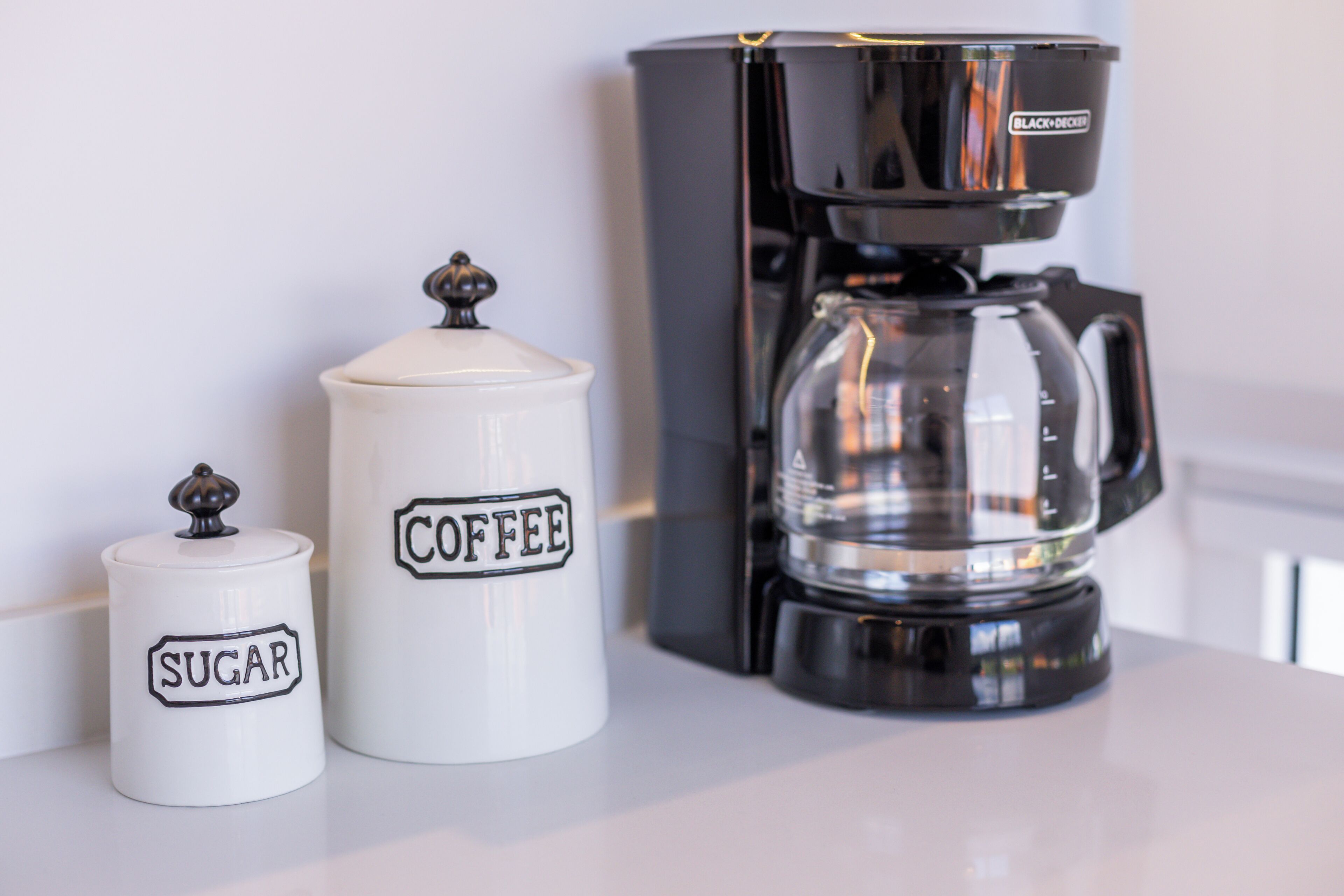 Two white kitchen containers labeled "SUGAR" and "COFFEE" with a black coffee maker beside them.