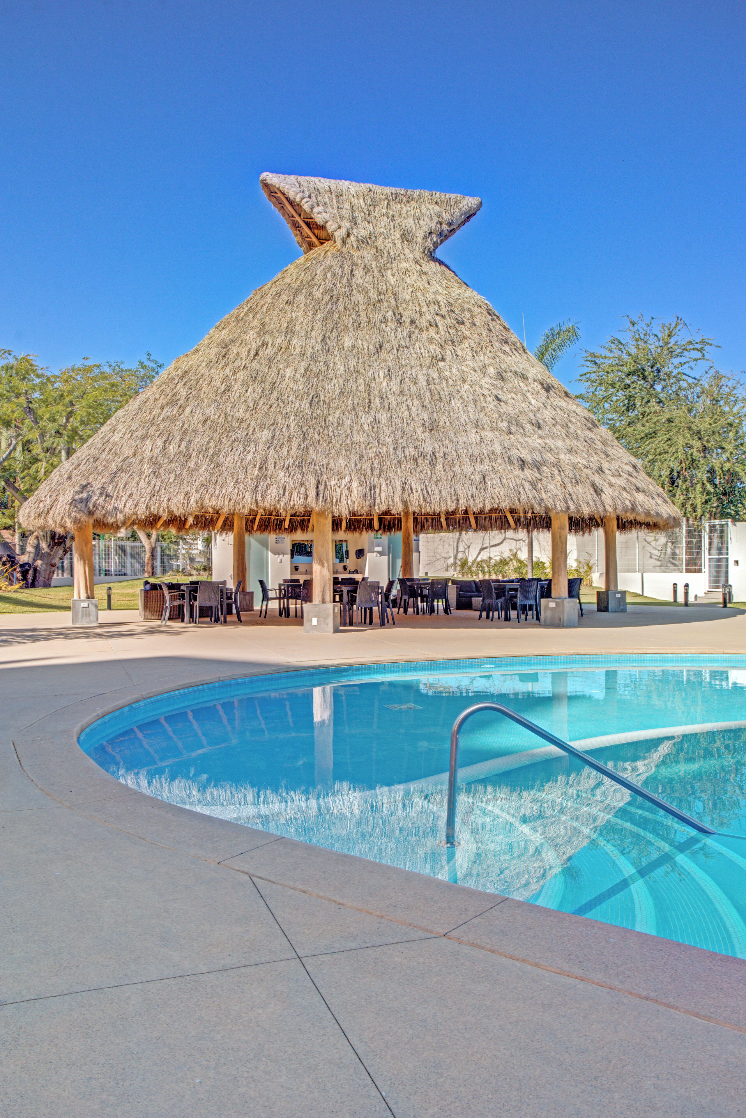 Un área de piscina con una estructura de techo de paja, sillas de descanso y una piscina azul cristalina.