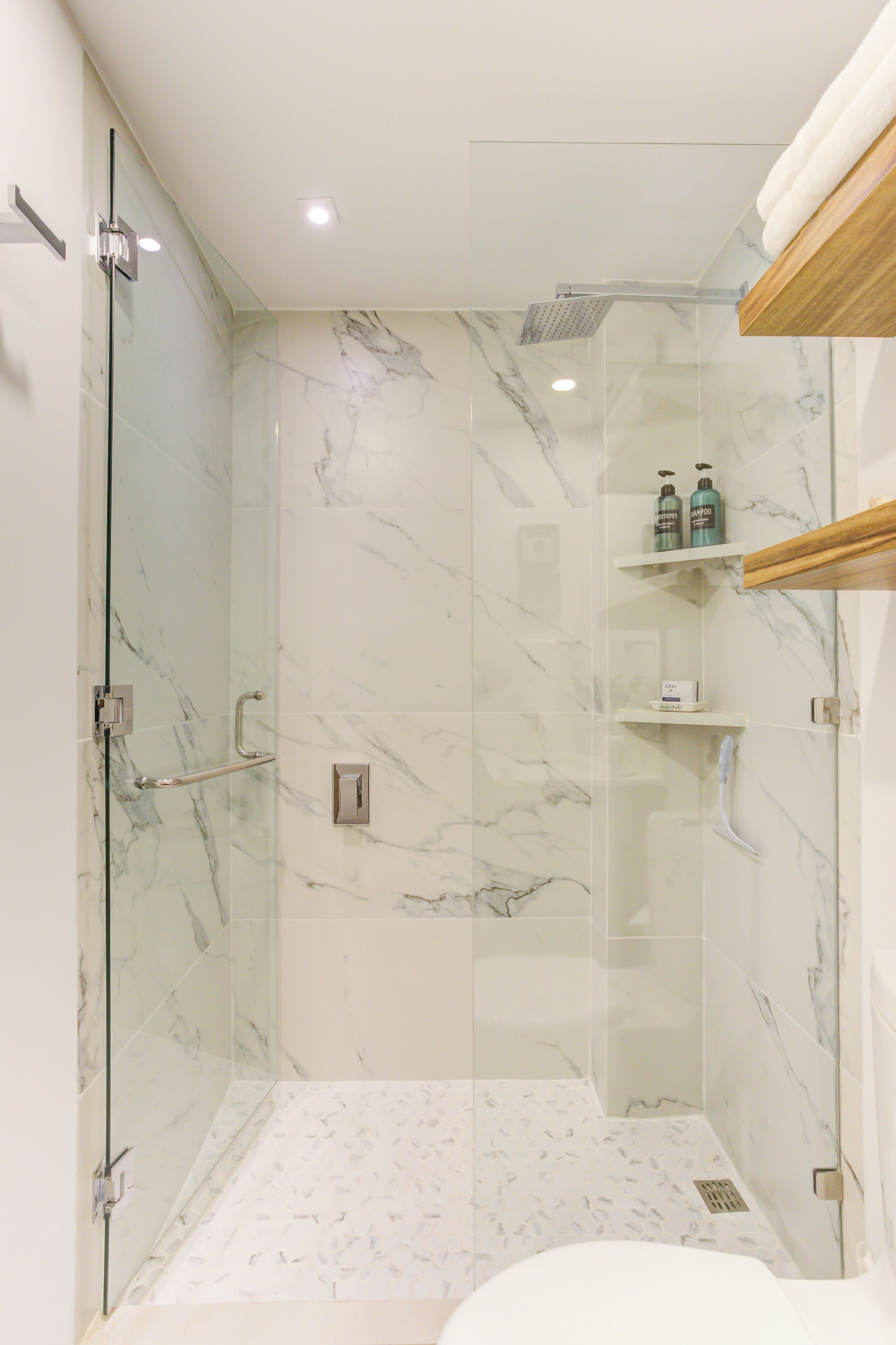 Un baño moderno con ducha de vidrio, baldosas de mármol y estante de madera con artículos de aseo.
