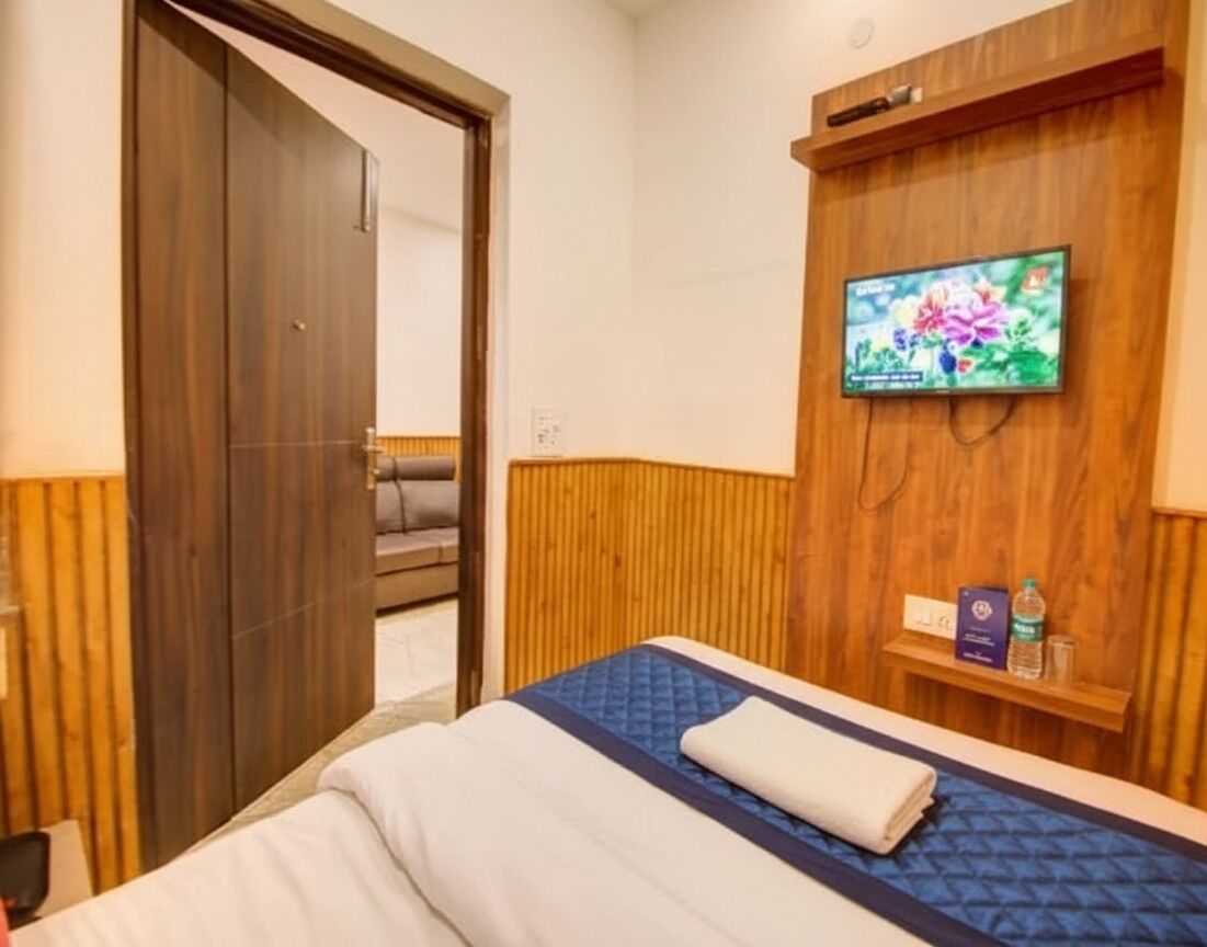Hotelkamer met een bed, een aan de muur bevestigde televisie, een houten wandpaneel en een klein plankje met een fles en een doos.