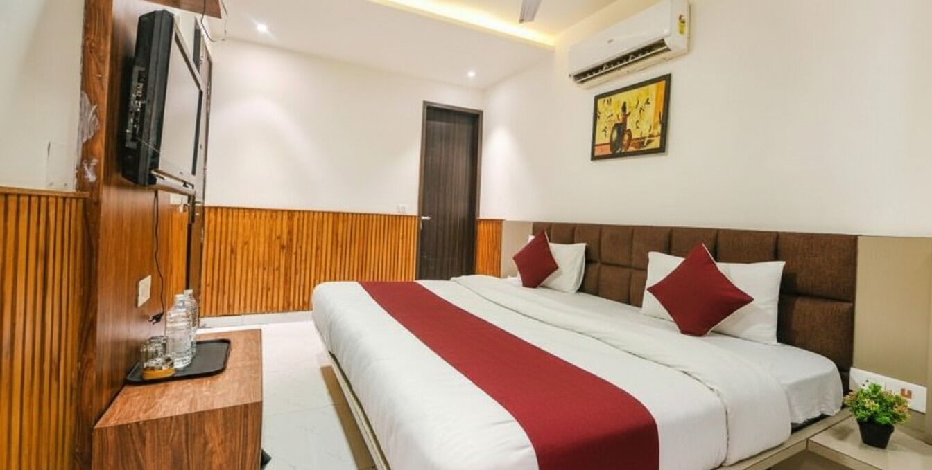 A hotel room with a bed, a wooden panel wall, a television, an air conditioning unit, and a small plant.