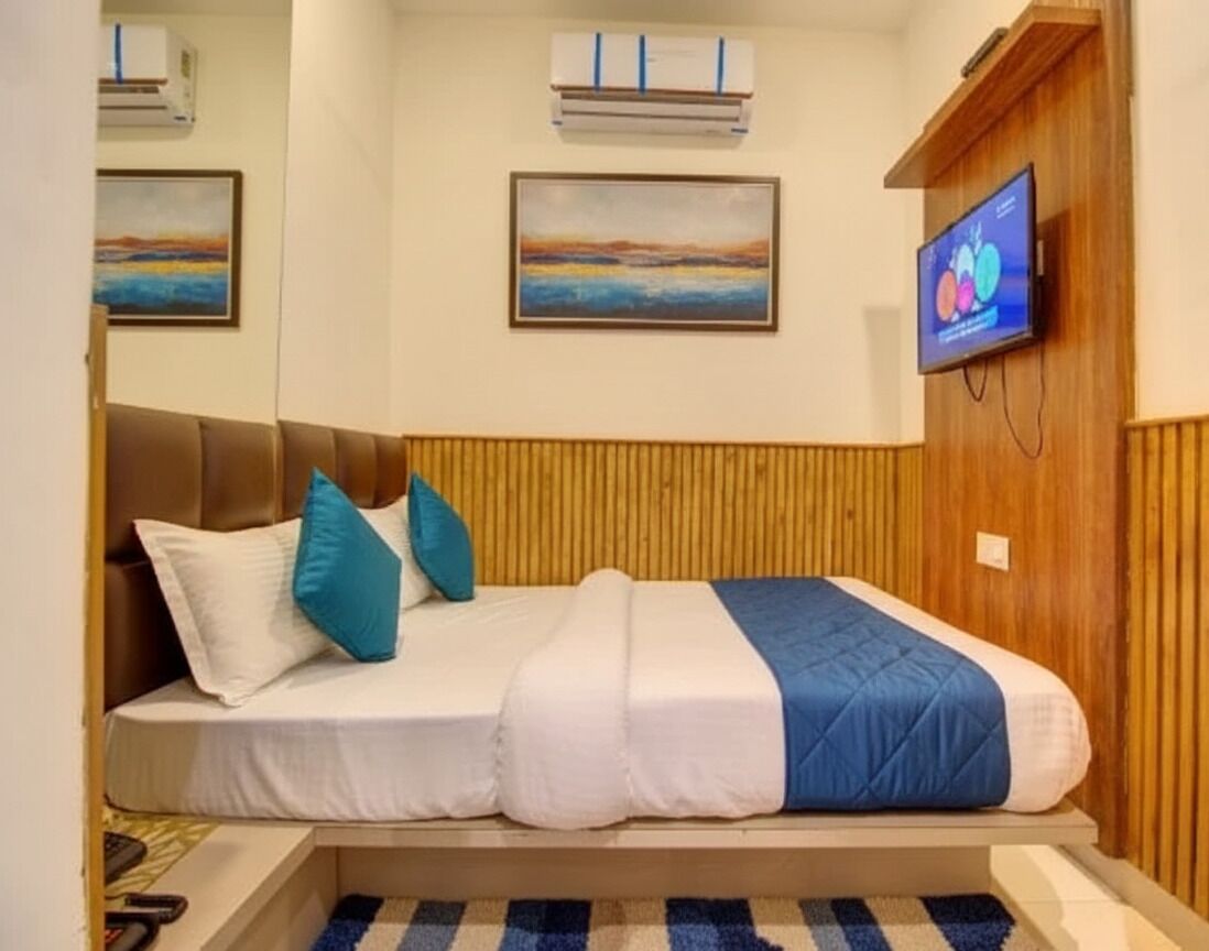 A single bed with a wooden headboard, white and blue bedding, and two pillows. There are two air conditioning units on the wall, a painting above the bed, and a television on the right.