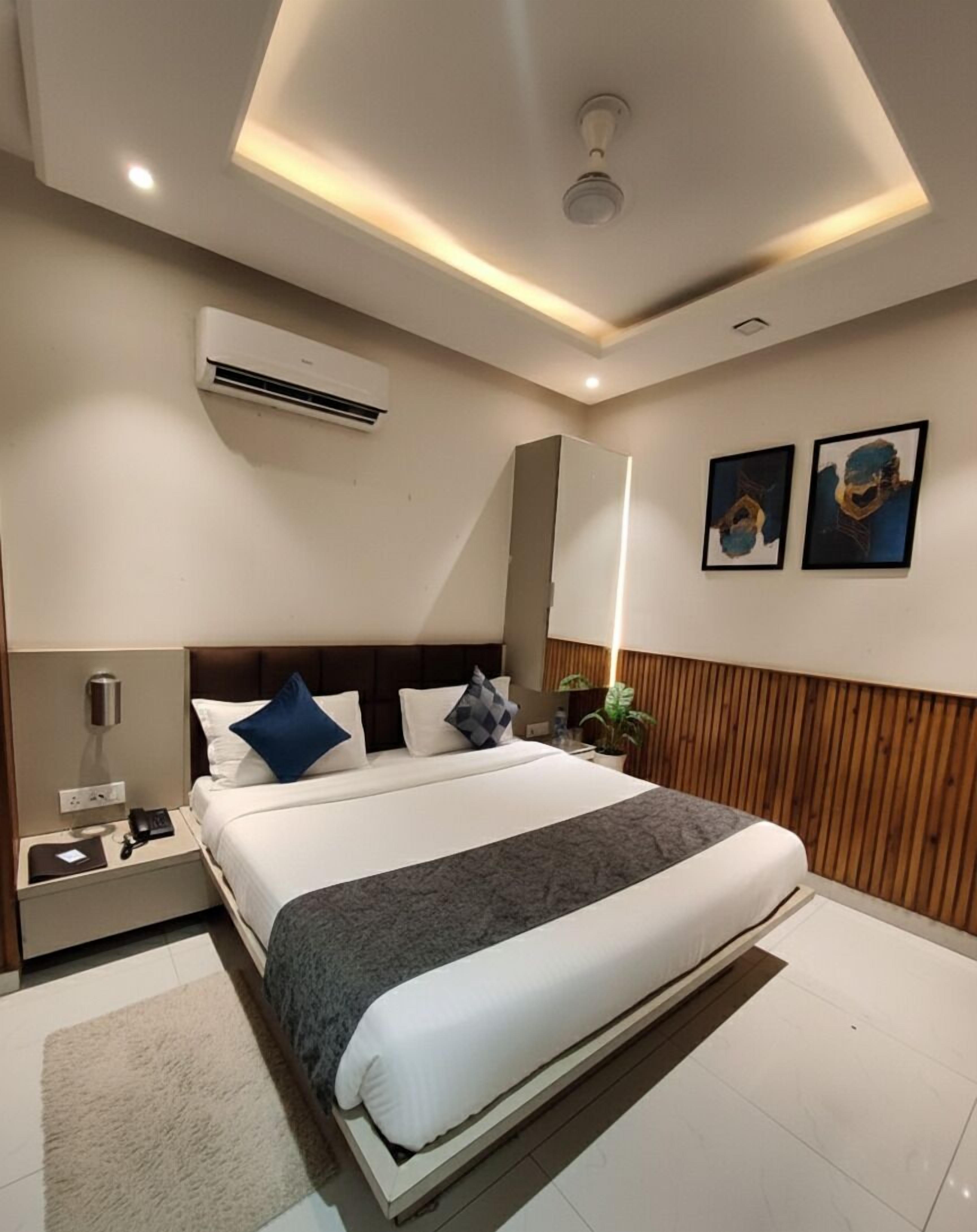 A modern hotel room with a large bed, a bedside table, an air conditioning unit, and two wall-mounted artworks.