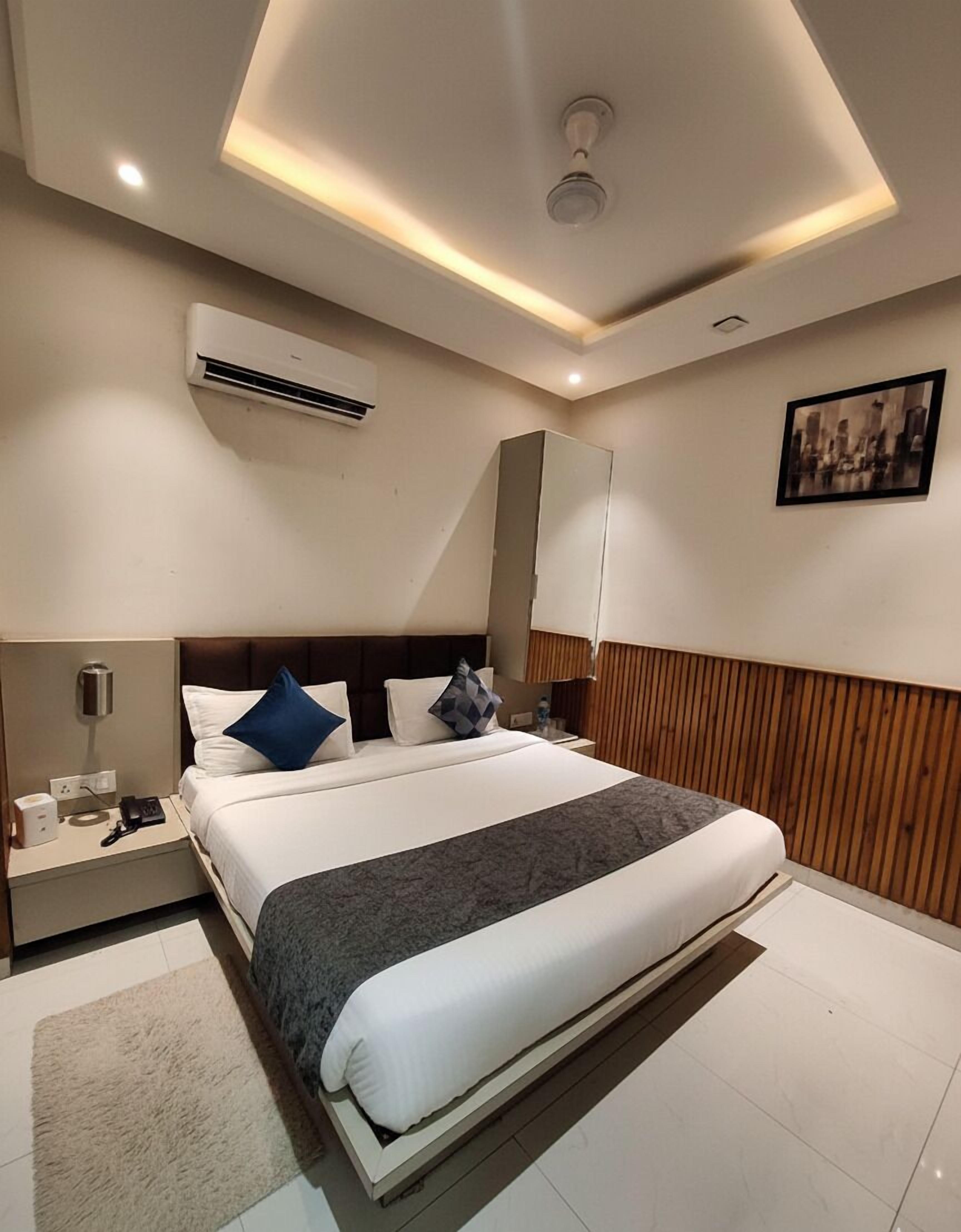 A modern hotel room with a large bed, a nightstand, an air conditioning unit, a wooden panel wall, and a cityscape painting.