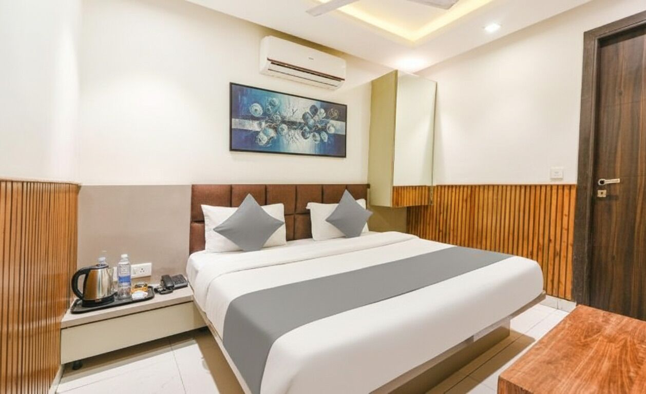 A modern hotel room with a large bed, a bedside table with a kettle, a water bottle, and a remote control. There is a painting on the wall above the bed and a wooden panel on the wall to the right.