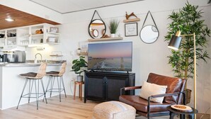 A modern living room with a sofa, a television, and a coffee table. There is a kitchen area with bar stools and a dining table. - 68th St Oceanfront Condo with Pool (Ocean City)