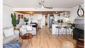 A modern living room with a sofa, coffee table, and a TV. There is a kitchen area with a fridge, microwave, and bar stools. - 68th St Oceanfront Condo with Pool (Ocean City)