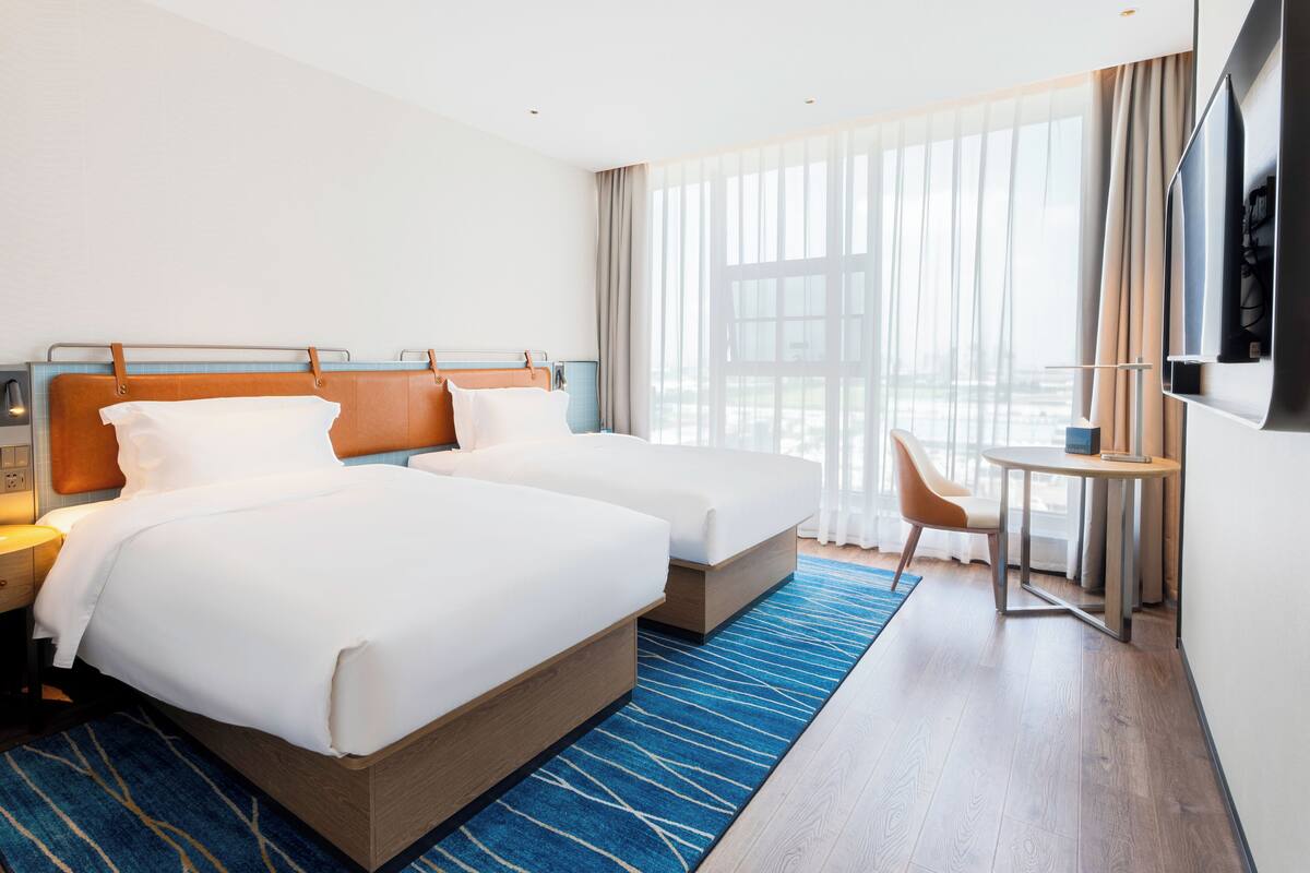 A hotel room with two beds, a desk, a chair, a TV, and large windows.