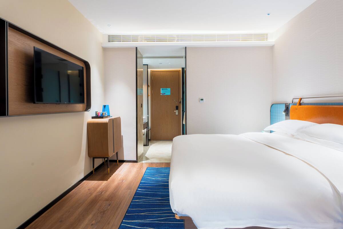 A modern hotel room with a large bed, a flat-screen TV mounted on the wall, a desk with a chair, and a blue patterned rug.