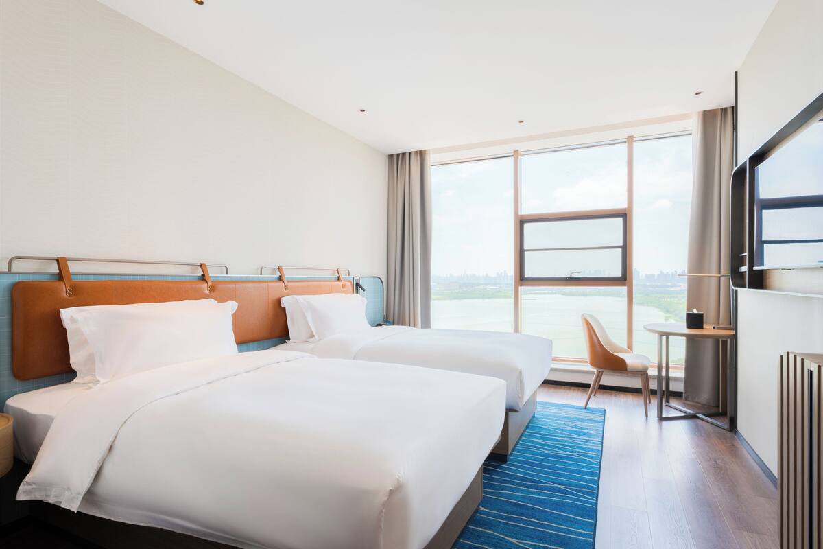 A hotel room with two beds, a large window with a city view, a small table, and a chair.