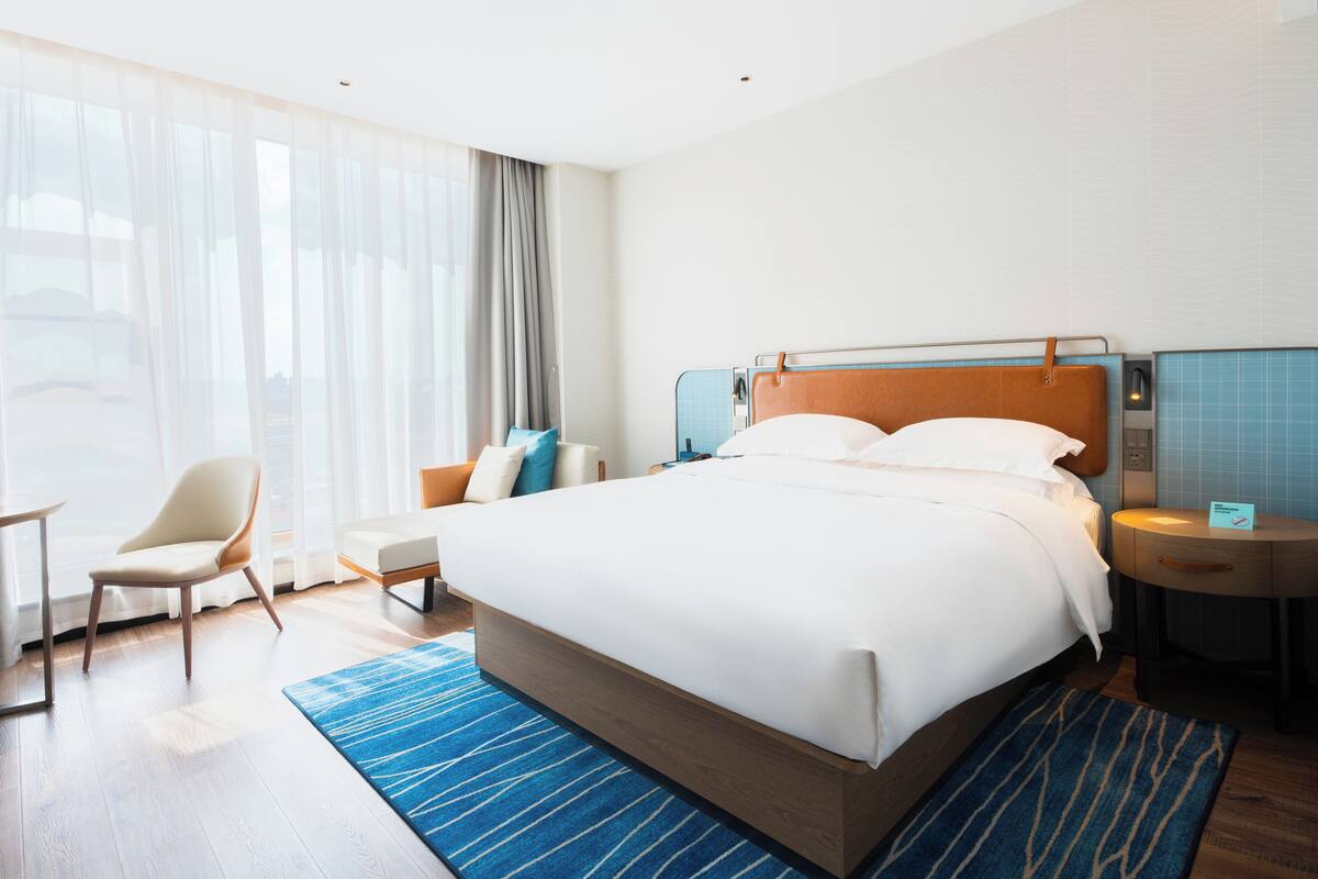 A modern hotel room with a large bed, a chair, a desk, and a view of the city through large windows.