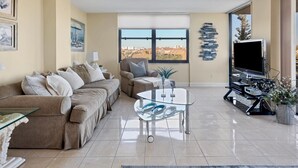 A living room with a beige sofa, a glass coffee table, a TV, and a large window with a city view.