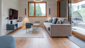 A modern living room with a sofa, two side tables, a TV, and a large window with a view of greenery. - Exclusive apartment right on the piste (Sonnenalpe Nassfeld)