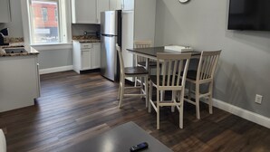 Une cuisine moderne avec des meubles blancs, un réfrigérateur en acier inoxydable, une table à manger avec des chaises et un téléviseur fixé au mur.