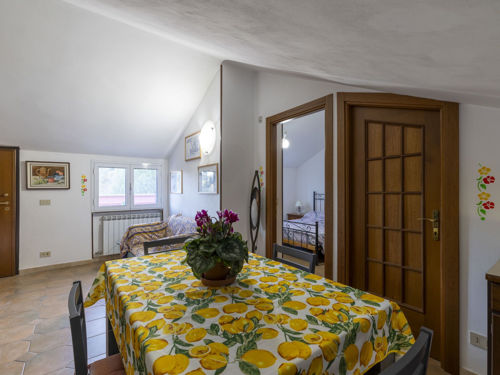 Ein Essbereich mit einem Tisch, der mit einem Zitronenmuster bedeckt ist, einer Topfpflanze und Blick auf ein Schlafzimmer durch eine offene Tür.