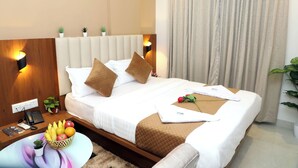 Una cama bien hecha con una combinación de colores blanco y marrón, una mesita de noche con una cesta de fruta, una planta pequeña y un mando a distancia.