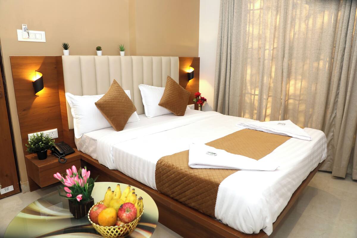 A neatly made bed with white linens, two pillows, and a brown throw blanket. There is a bedside table with a lamp, a phone, and a small plant. A fruit basket with bananas, apples, and oranges is on the floor beside the bed.