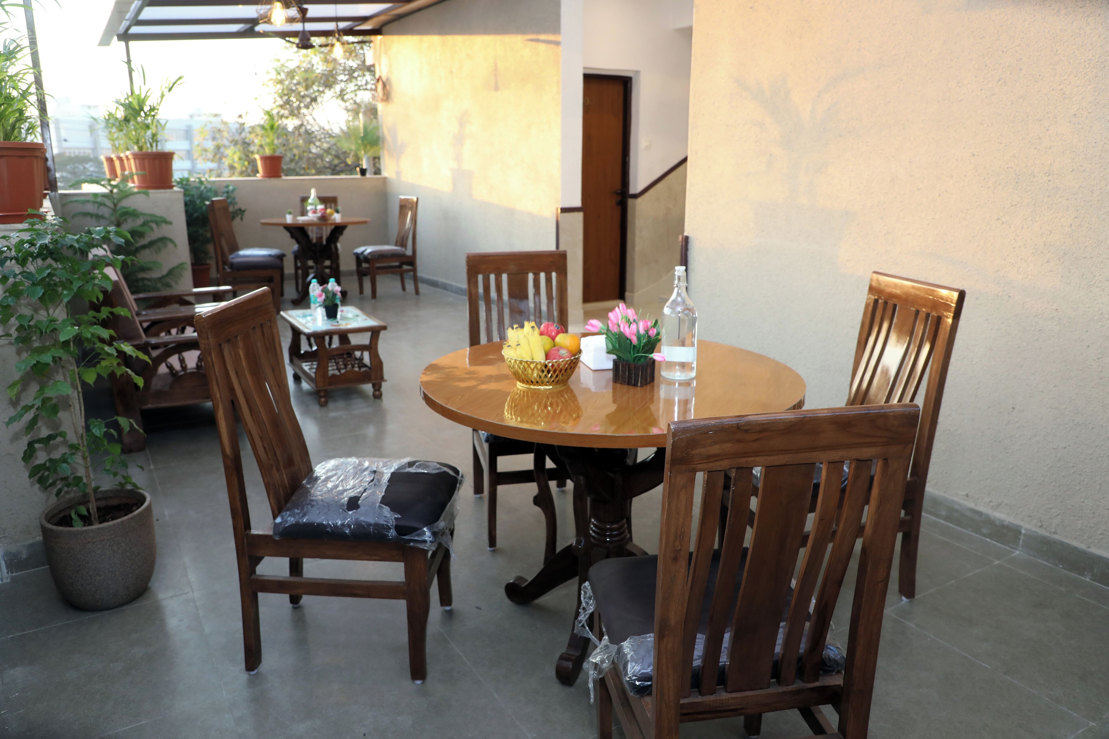 A round wooden table with chairs, surrounded by potted plants and a small table with a basket of fruit and flowers.