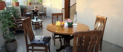A round wooden table with chairs, surrounded by potted plants and a small table with a basket of fruit and flowers.