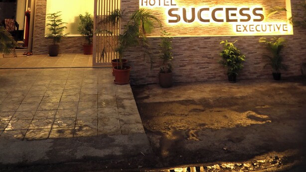 Un hotel llamado "Hotel Success Executive" con una fachada bien iluminada, ventanas amplias y plantas en macetas alrededor de la entrada.