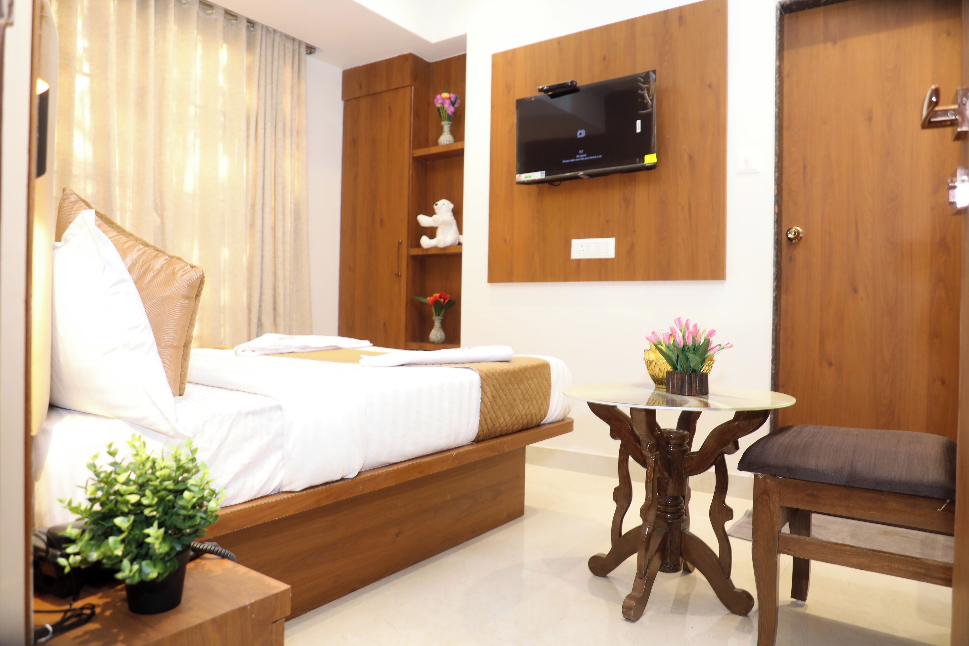 A hotel room with a bed, bedside table, TV mounted on the wall, a wooden door, and a small table with a chair.
