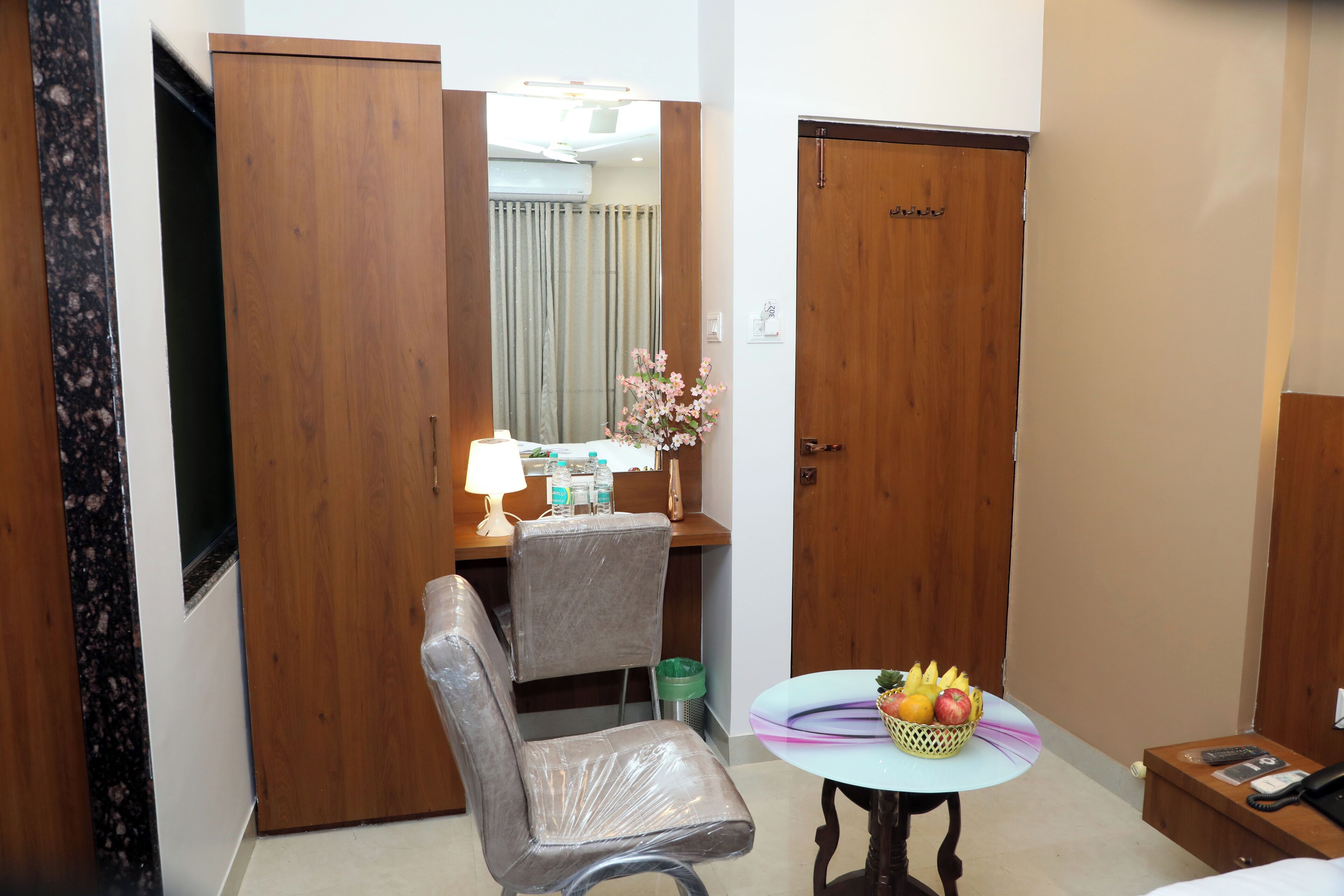 A hotel room with a wooden wardrobe, a desk with a lamp, a chair, a small table with fruit, and a bed.