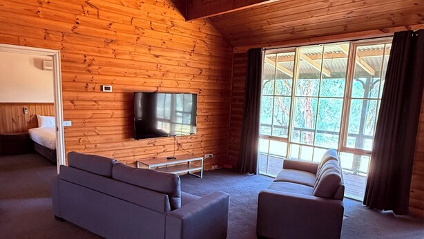 Interior d'una cabana de fusta amb una finestra gran, un televisor de pantalla plana i dos sofàs.