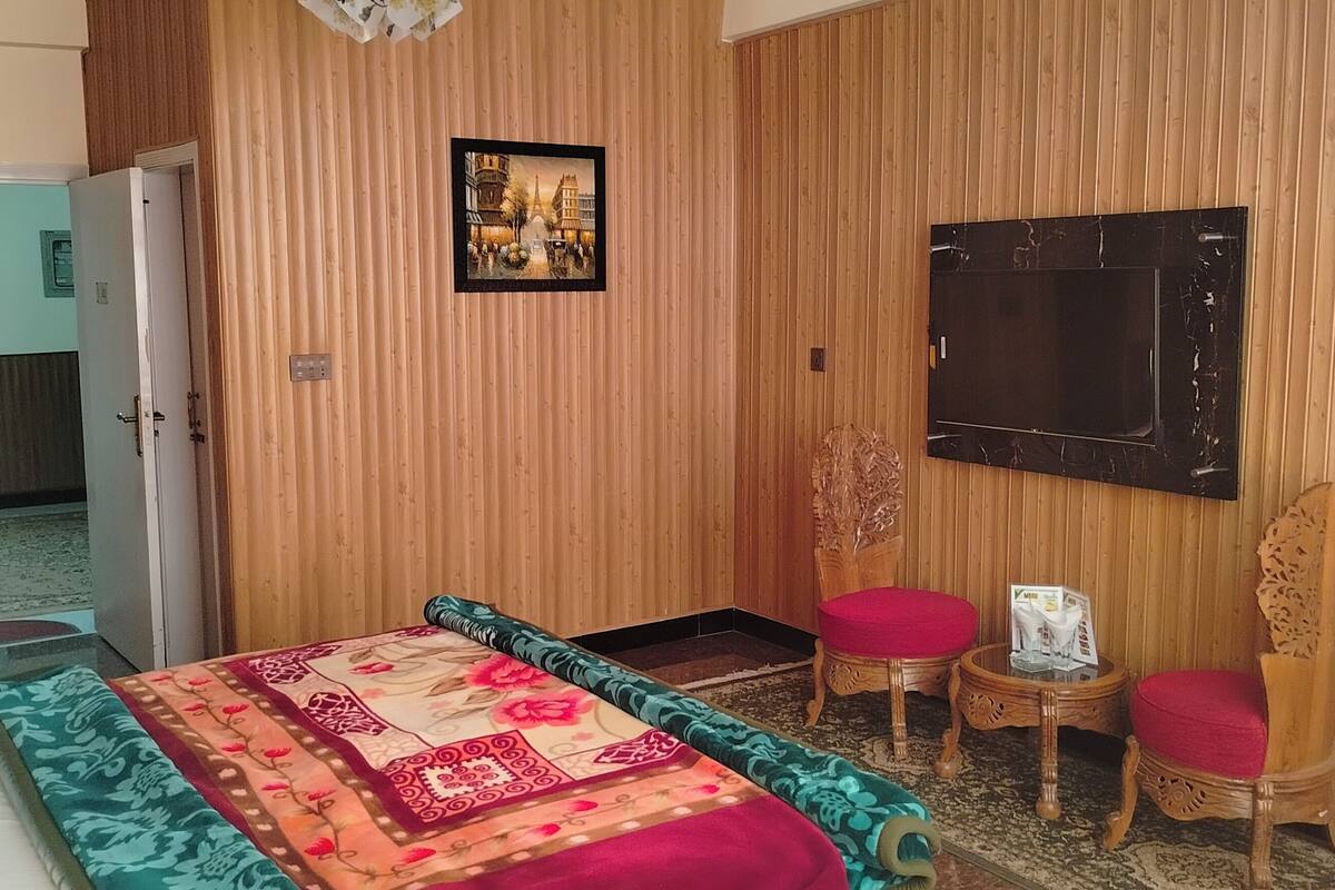 Quarto de hotel com uma cama, duas poltronas vermelhas com almofadas, uma mesa pequena com uma revista, um quadro na parede e uma televisão fixada na parede.