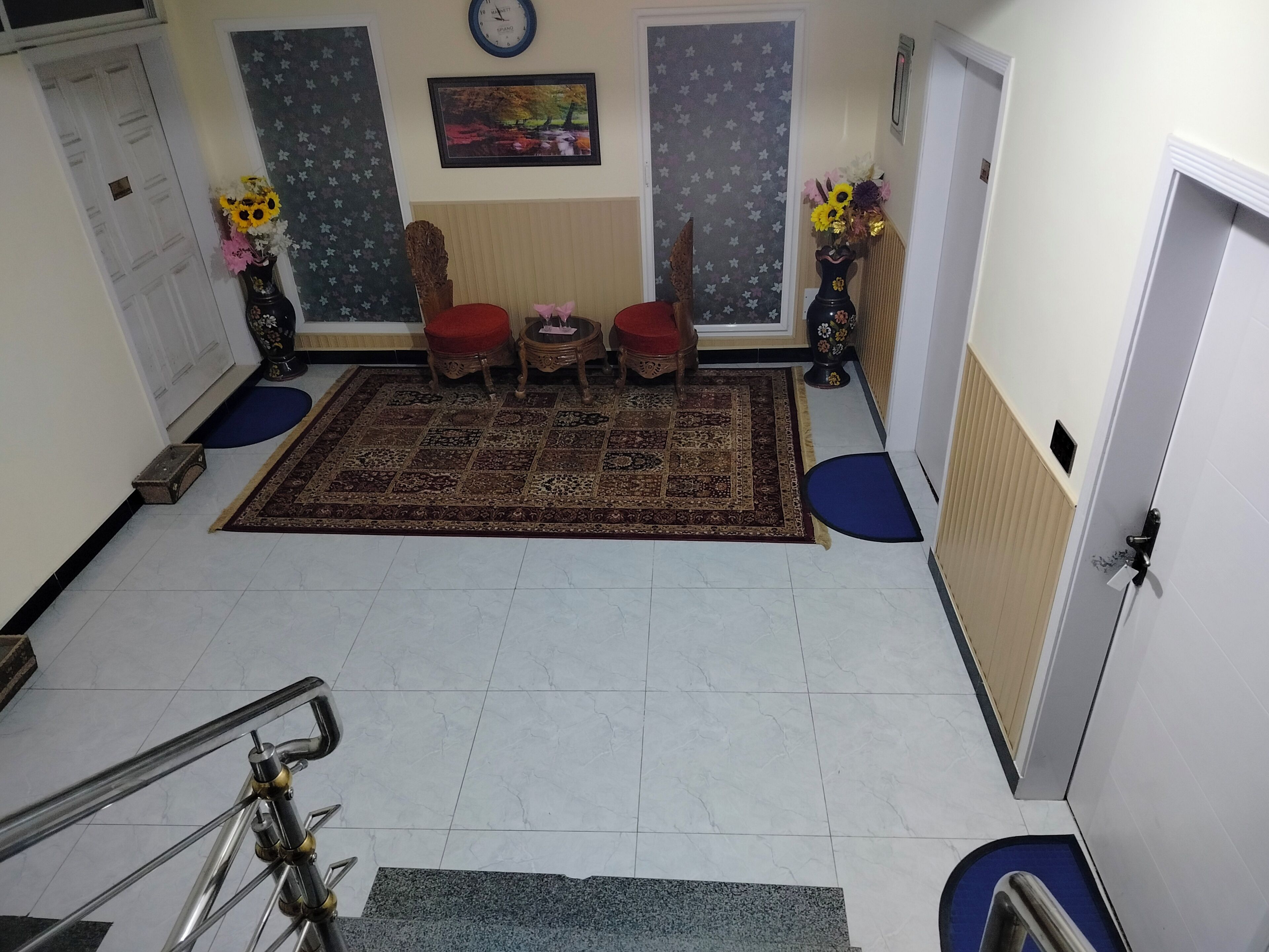 Un amplio salón con un tapete estampado, dos sillones rojos, una mesita y dos mesitas de noche con jarrones florales.