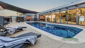 Un área de piscina moderna con sillones de descanso, un edificio con paredes de vidrio y un cielo despejado.